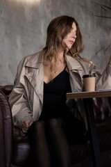 Fototapeta na wymiar beautiful attractive slim brunette in trench coat in a grey loft room on leather couch with a paper cup. urban lifestyle concept. 