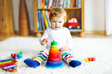Adorable cute beautiful little baby girl playing with educational toys at home or nursery. Happy healthy child having fun with colorful wooden rainboy toy pyramid. Kid learning different skills
