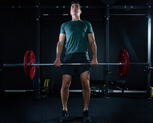 Tall dark man with green t shirt lifting barbell with red weights on a dark gym.