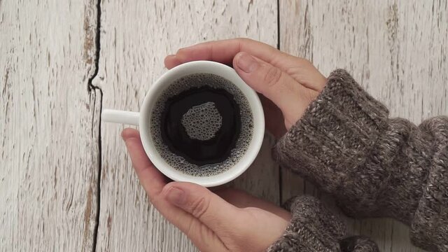 Woman In Sweater Holding A Cup Of Coffee In Her Hands. Brown Drink With Foam Rotating After Stirring. Top Down Shot.