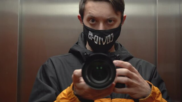 Portrait of one young brunette man in elevator. Guy wearing black, yellow jacket, protective face mask with inscription Covid-19, holds mirrorless camera, takes pictures of himself in mirror close up
