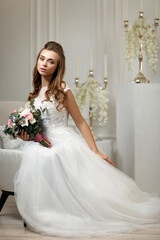 beautiful tender young bride in a luxurious wedding dress with flower bouquet sitting on armchair in white studio interior