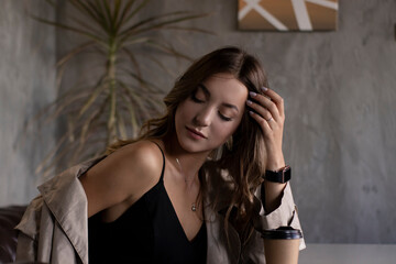 Fototapeta premium beautiful attractive slim brunette in trench coat in a grey loft room on leather couch with a paper cup. urban lifestyle concept. 