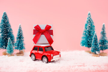 Miniature red car toy delivering gift box and christmas tree on pink background. Christmas greeting card concept.