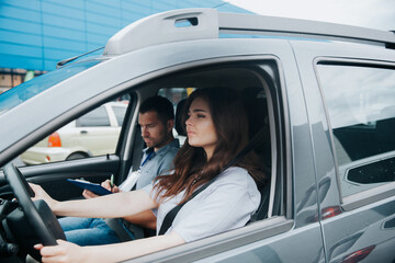 Young upset woman takes driving exam with serious male auto instructor. Two people sitting in gray car with fastened seat belts. Concentrated young woman holds steering wheel and looks forward.