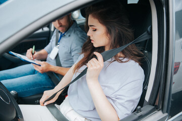 Beautiful young woman upset with results of driving tests. Strict examiner holds clipboard and makes notes of her mistakes. Female student and male instructor sit in a car with open window.