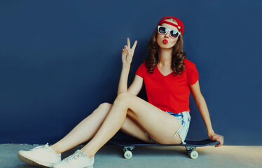 Attractive woman sitting on skateboard showing peace sign gesture wearing baseball cap over blue...
