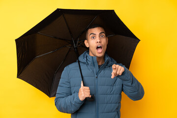 Young handsome man holding an umbrella over isolated yellow background surprised and pointing front