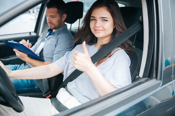 Female student showing thumbs up and smiling. Cheerful young woman excited about her successful driving license exam. Male driving instructor wrtiting something in his paper. Safe driving concept.