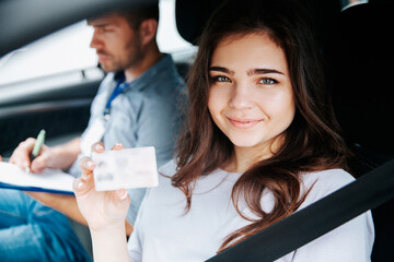 Attractive young woman showing driver's licence, looking at camera and smiling. Brunette woman sitting on drivers seat with fastened seat belt. Blurred male instructor on background writing something.