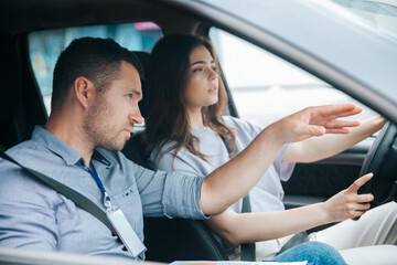 Male instructor showing something by his hand on the road and giving advice. Attractive woman listening her teacher, looking ahead with concentration and holding wheel.