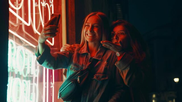 Happy flirting friends, send love and kisses to followers on social media. Two young women pose for selfie photos or video on smartphone camera. Night time screen lit image in neon lights