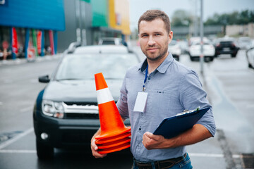 Male driving instructor standing with orange traffic cones and clipboard in his hands. Caucasian man working in driving school. Blurred gray car in background. - obrazy, fototapety, plakaty