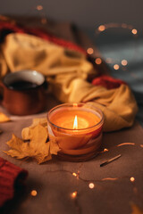 Burning aroma candle with autumn leaves and light garland decor on brown plaid in bed. Comfort and relaxation concept.