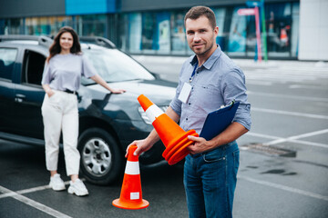 Happy male driving instructor holding traffic cones in his hands, looking at camera and smiling. Young woman standing near the car and waiting when man finish his job.