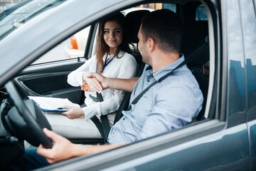 Female auto instructor shaking hands with male student. Attractive woman congratulates with successful driving license exam. Man in blue shirt holding wheel by his hand and looking at woman.