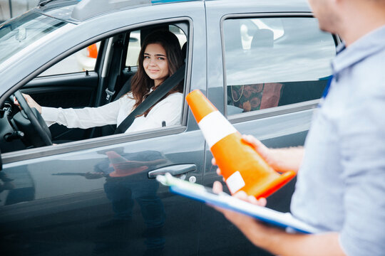 Positive experience in driving school. Cheerful confident young woman is glad for improvement of her driving skills. Male instructor stands near the car holding orange road cone and clipboard.