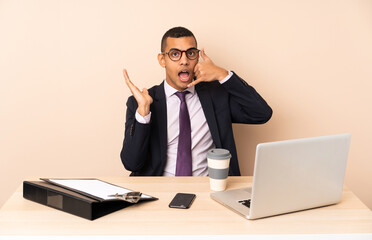 Young business man in his office with a laptop and other documents making phone gesture and doubting