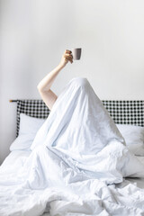Fototapeta na wymiar Young woman with cup of coffee or tea sitting in bed under blanket at bedroom. Staying at home
