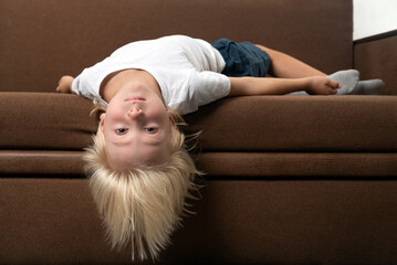 Child lies on the couch with his head hanging down. Boy is tired of lessons