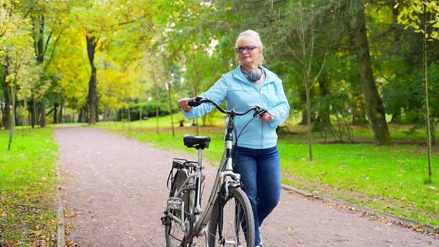 Happy senior woman in glasses casual clothes walks in the park with a bicycle. old female cyclist with a retro bike pursues active lifestyle in nature. A walk in the autumn forest or city park outside