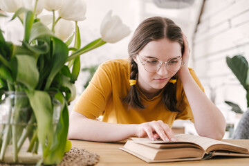 Young beautiful woman student in orange T-shirt and glasses reads book sitting at the table....