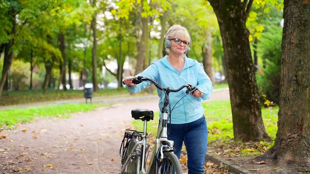 happy senior woman in headphones walks in the park with a bicycle and listens music audio book. old female cyclist with retro bike pursues an active lifestyle in nature. autumn forest or city outside