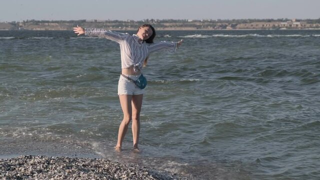 Happy young caucasian woman stands on seashore and opens her arms to sides. Girl enjoying sunny day on beach. Wind blows femals hair. Carefree seaside vacation. Wide shot. Slow motion