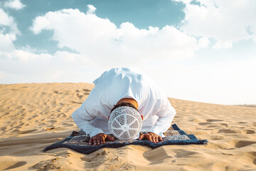 Man with white traditional kandura from uae praying in the desert on the carpet