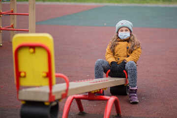 Little girl in medical mask on seesaw. At atumn playground