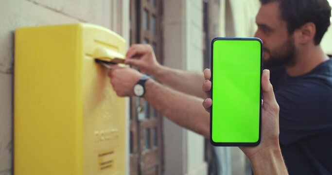 Close up view of female hand holding smartphone with mock up screen while bearded man putting letter in post box. Concept of greenscreen and chroma key. Outdoors.