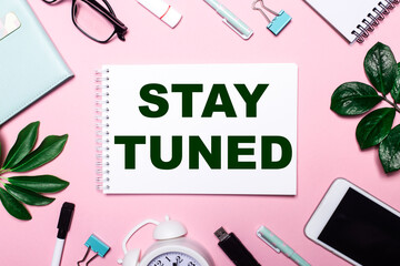 STAY TUNED is written in a white notebook on a pink background surrounded by business accessories and green leaves.