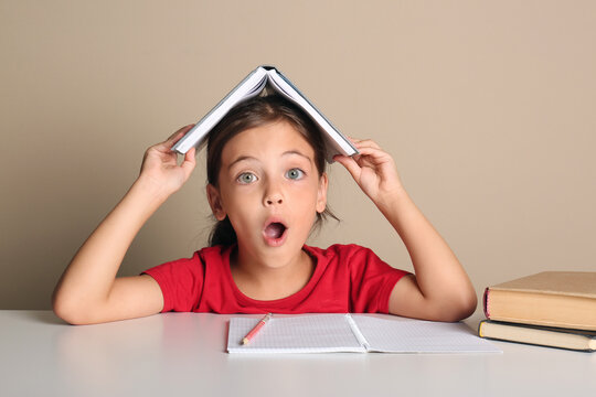 Emotional little girl with book on her head doing homework at table against beige background