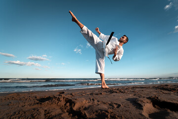 Black belt karate fighter giving a high kick on the beach at sunset