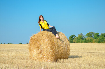 Naklejka na ściany i meble Young redhaired slender woman of european appearance in black and yellow clothes sits on haystack on field under sun