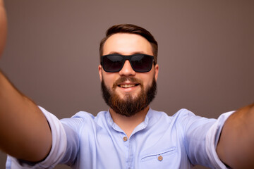 Close up of cheerful young bearded man in classic blue shirt and sunglasses make selfie isolated on grey  background. 