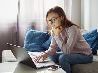 Young woman with glasses sitting on the couch and working on a laptop. A female student is...