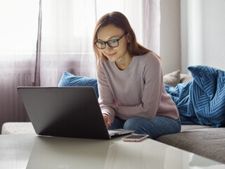 Smiling woman working on a laptop. Young, satisfied girl with glasses sitting on the couch in front of the laptop. There's a smartphone on the table.