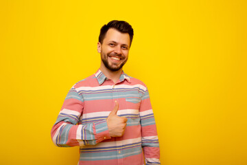 Young handsome man with beard wearing casual shirt feeling happy and positive, showing thumb up doing excellent and approval sign. He standing isolated on yellow background. 