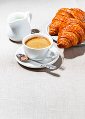 Croissants and cup of coffee in the morning light, sunlight shadow, on a linen fabric. Morning breakfast concept.