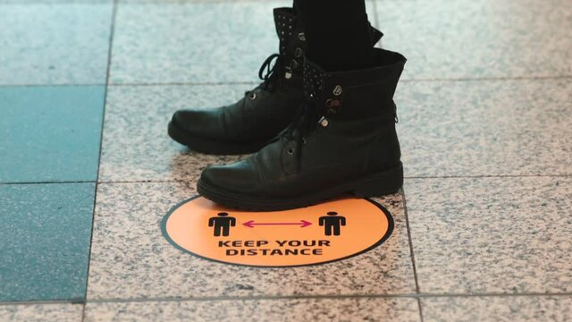 Legs in black boots stand at a orange "keep your distance" sign in a public place due to the need to maintain a physical distance for safety reasons. The new normality rules. 