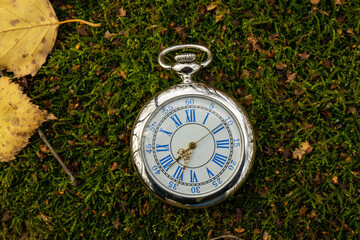 Vintage pocket watch on the last green grass surrounded by yellow autumn leaves, Close-up.