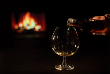 Woman pours cognac into a glass on the background of the fireplace.
