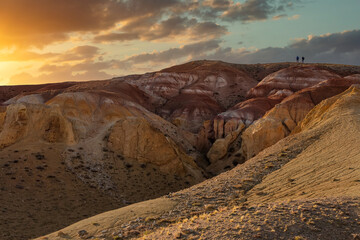 Fototapeta na wymiar Beautiful low angle shot of two tiny human figures climbing massive red mountain in Kyzyl-Chin valley, also called Mars valley. Sunset cloudy sky as a backdrop. Golden hour. Altai, Siberia, Russia