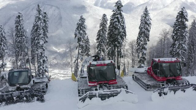 Snow groomers standing on parking lot in snowy mountain at winter ski resort. Aerial view red machine on winter highlands on snowy forest landscape in resort