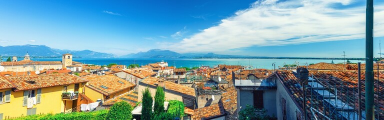 Naklejka na ściany i meble Panorama of Desenzano del Garda town with red tiled roof buildings, Garda Lake water, Monte Baldo mountain range, Sirmione peninsula, Lombardy, Northern Italy. Aerial panoramic view of Desenzano