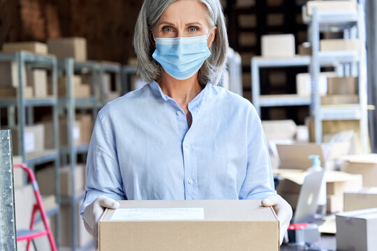 Older Business Woman Retail Seller, Entrepreneur, Small Business Owner Wears Face Mask And Gloves For Covid 19 Protection Looking At Camera Holding Parcel Box Standing In Delivery Warehouse, Portrait.