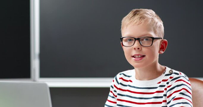 Portrait of small teen Caucasian boy in glasses ooking at camera and sitting at school. Indoor. Blackboard on background. Close up of face of teenage schoolboy in eyeglasses in classroom.
