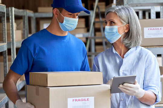 Female manager supervisor wearing face mask using digital tablet talking to male courier holding shipping parcels boxes discuss delivery walking in warehouse. Covid 19 safety social distance at work.