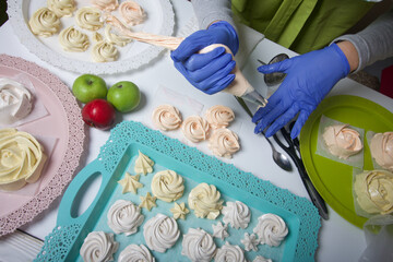 A woman in rubber gloves and a medical mask makes homemade marshmallows. Fresh marshmallows are nearby on a tray. Cooking sweets during a pandemic.
